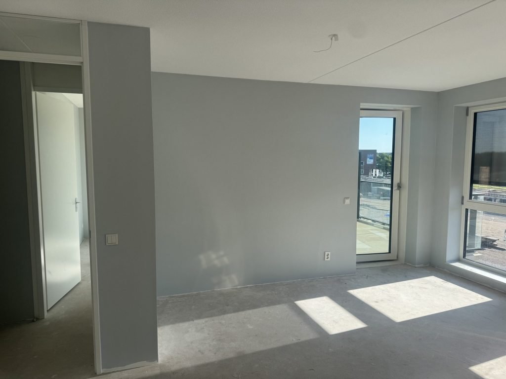 Lege nieuwbouwkamer met kale wanden, een betonvloer en een balkondeur, kort na de oplevering
